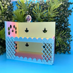 May include: A decorative, multi-tiered shelf in pastel colors. The shelf has a light blue scalloped edge and a lattice design on the sides. The interior is a gradient of pink, yellow, and orange. The background is a vibrant blue with green foliage.