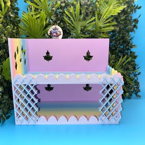 May include: A two-tier pastel shelf featuring a light purple back panel with leaf-shaped cutouts, a rainbow gradient base, and light blue lattice-style side panels. The front edges of the shelves have a scalloped design, creating a decorative display unit.