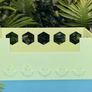 May include: A light green rectangular organizer with a series of hexagonal cutouts and leaf-shaped designs. The organizer is made of a solid material and is designed to hold small items. The background is a mix of green foliage and a blue surface.
