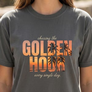 以下が含まれることがあります： 「chasing the GOLDEN HOUR every single day.」というテキストがプリントされたチャコールグレーの半袖Tシャツ。「GOLDEN HOUR」の文字の中には、ヤシの木、オレンジ色の空、海の波が描かれた夕暮れのビーチの風景がデザインされています。