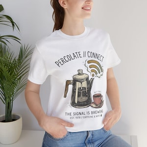 May include: White t-shirt with a coffee-themed graphic. The design features a vintage coffee pot, a coffee cup, and a WiFi symbol. Text on the shirt reads "PERCOLATE // CONNECT" and "THE SIGNAL IS BREWING EST. 1978 / CAFFEINE & WIFI".