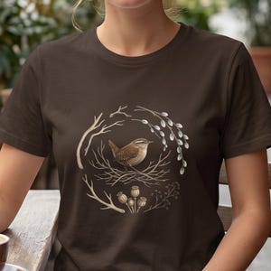 May include: A dark brown short-sleeved t-shirt featuring a central graphic of a small brown bird perched in a nest. The bird is surrounded by a circular arrangement of bare twigs, pussy willow branches, and dried poppy seed pods in neutral tones.