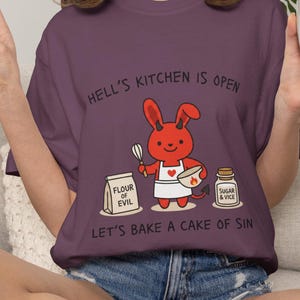 May include: A purple t-shirt with the text "HELL'S KITCHEN IS OPEN" and "LET'S BAKE A CAKE OF SIN". The graphic features a red devil bunny with a whisk and bowl, and bags labeled "FLOUR OF EVIL" and "SUGAR & VICE".