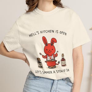 May include: Cream-colored t-shirt with the text "HELL'S KITCHEN IS OPEN" and "LET'S SIMMER A STEW OF SIN." A cartoon devil bunny is cooking in a pot. Two bottles and a jar are also visible.