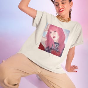 May include: Cream-colored t-shirt featuring a portrait of a woman with pink hair. The artwork is on a red and blue background. The person in the image is wearing tan pants, white sneakers, and a gold chain necklace.