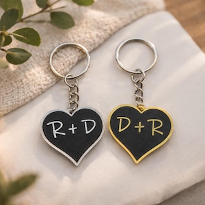 Könnte beinhalten: Zwei herzförmige Schlüsselanhänger mit Initialen. Einer hat einen silbernen Rand und die Initialen "R+D" in Weiß auf schwarzem Hintergrund. Der andere hat einen goldenen Rand und die Initialen "D+R" in Gelb auf schwarzem Hintergrund.