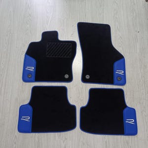 Könnte beinhalten: Ein Set aus vier schwarzen Auto-Fußmatten mit blauem Rand und weiß gestickten R-Logos. Die Matten verfügen über eine strukturierte Fersenauflage auf der Fahrerseite sowie kreisförmige Befestigungsösen für den Einbau im Fahrzeuginnenraum.