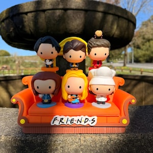 Könnte beinhalten: Ein Set von Figuren, die Charaktere aus der Fernsehserie "Friends" darstellen. Die Figuren sind auf einem orangefarbenen Sofa angeordnet, mit dem Logo der Serie. Jede Figur hat eine eigene Kleidung und Frisur. Der Hintergrund umfasst eine Steinmauer und einen Brunnen.