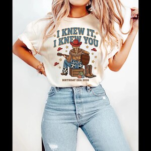 May include: A cream-colored t-shirt featuring a graphic of a person in a red cowboy hat, brown fringe jacket, and cow-print jeans sitting on a wooden crate playing a guitar. The text reads I KNEW IT, I KNEW YOU and BIRTHDAY ERA 2026.