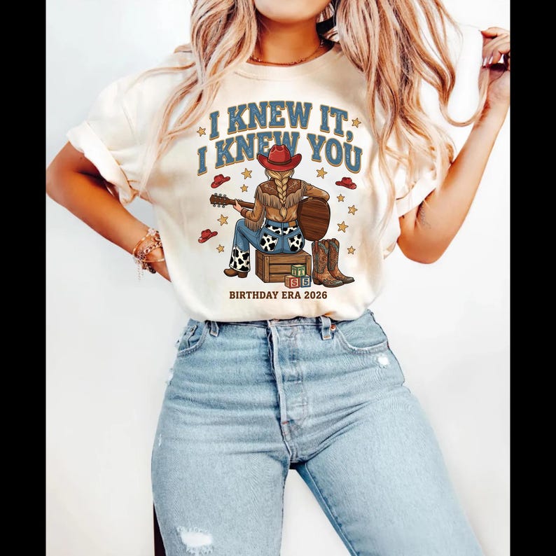 May include: A cream-colored t-shirt featuring a graphic of a person in a red cowboy hat, brown fringe jacket, and cow-print jeans sitting on a wooden crate playing a guitar. The text reads I KNEW IT, I KNEW YOU and BIRTHDAY ERA 2026.