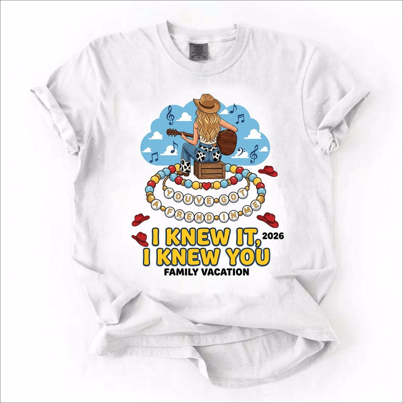 May include: A white t-shirt featuring a graphic of a person in a cowboy hat playing a guitar, sitting on a crate. Below are beaded necklaces with the text "YOU'VE GOT A FREND IN ME" and the printed text "I KNEW IT, I KNEW YOU 2026 FAMILY VACATION".
