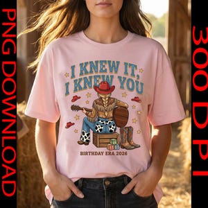 May include: A light pink t-shirt featuring a graphic of a person with a blonde braid wearing a red cowboy hat, fringe jacket, and cow-print jeans, sitting on a crate playing a guitar. Text reads I KNEW IT, I KNEW YOU and BIRTHDAY ERA 2026.
