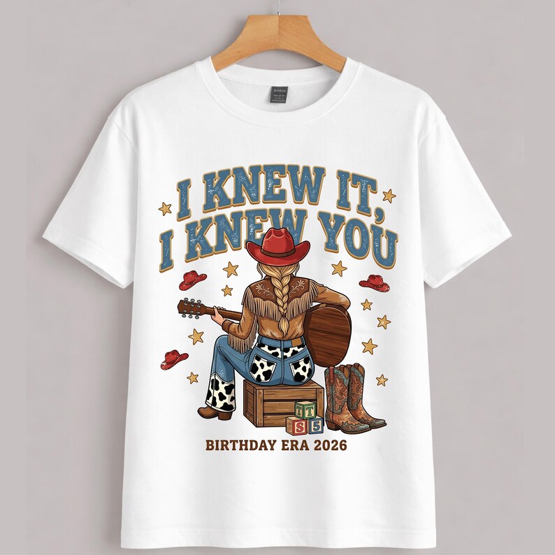May include: A white short-sleeve t-shirt featuring a graphic of a person with a braided hairstyle wearing a red cowboy hat, a brown fringed jacket, and cow-print jeans, playing a guitar while sitting on a wooden crate with blocks labeled L, I, S, 5.