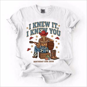 May include: A white t-shirt featuring a graphic of a person with a blonde braid wearing a red cowboy hat, fringe jacket, and cow-print pants, sitting on a wooden crate playing a guitar. Text reads I KNEW IT, I KNEW YOU and BIRTHDAY ERA 2026.