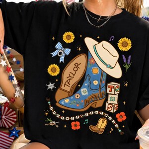 May include: A black t-shirt featuring a graphic print of a blue cowboy boot with fringe, a beige cowboy hat, sunflowers, a blue gingham bow, and blocks labeled T, 13, and S. The text "TAYLOR" and "I KNEW IT WHEN YOU" are printed on the shirt.