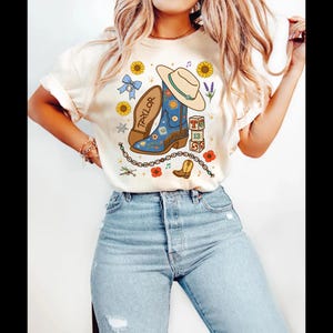May include: A cream-colored t-shirt featuring a graphic print of a blue cowboy boot, a beige cowboy hat, sunflowers, a blue gingham bow, alphabet blocks spelling TS 13, a beaded friendship bracelet, and the name Taylor written on a boot sole.