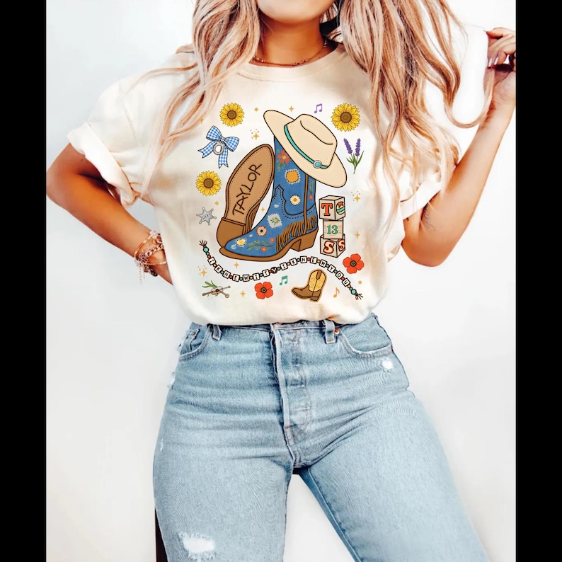 May include: A cream-colored t-shirt featuring a graphic print of a blue cowboy boot, a beige cowboy hat, sunflowers, a blue gingham bow, alphabet blocks spelling TS 13, a beaded friendship bracelet, and the name Taylor written on a boot sole.