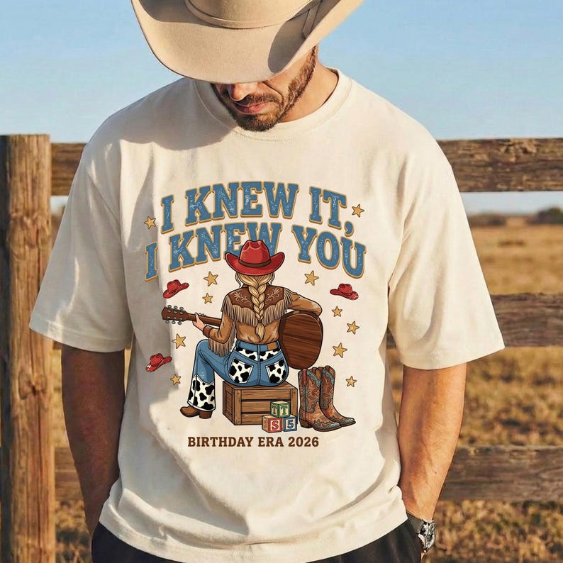 May include: A cream-colored t-shirt featuring a graphic of a person in a red cowboy hat and cow-print pants playing an acoustic guitar while sitting on a wooden crate. The text reads I KNEW IT, I KNEW YOU and BIRTHDAY ERA 2026 with stars.