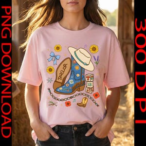 May include: A light pink t-shirt featuring a graphic print of a blue cowboy boot, a white cowboy hat, a blue checkered bow, sunflowers, and a stack of blocks with the text 13. The design includes a beaded string and the name Taylor.
