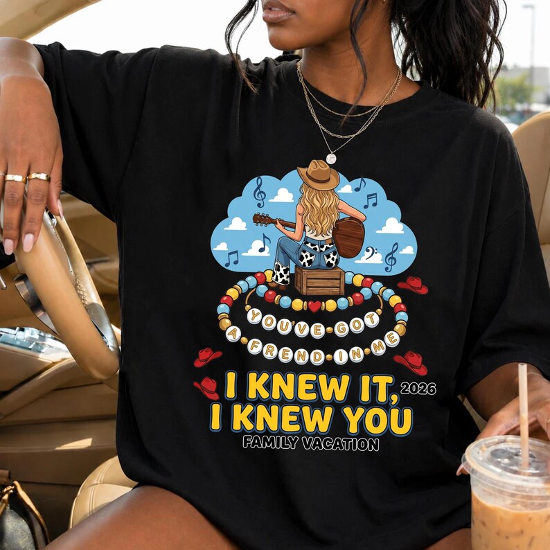 May include: A black t-shirt featuring a graphic of a blonde person in a cowboy hat playing a guitar while sitting on a crate. Below, a beaded necklace spells out YOU'VE GOT A FREND IN ME, followed by the text I KNEW IT, I KNEW YOU 2026 FAMILY VACATION.