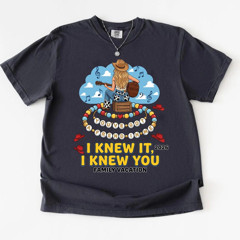 May include: A dark charcoal gray t-shirt featuring a graphic of a person in a cowboy hat playing a guitar while sitting on a crate. The design includes a beaded necklace with the text YOU'VE GOT A FREND IN ME, and the text I KNEW IT, I KNEW YOU 2026 FAMILY VACATION.