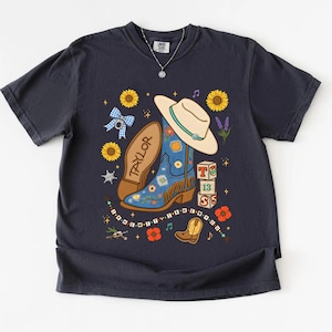 May include: A dark charcoal gray t-shirt featuring a graphic print of a blue cowboy boot, a beige hat, sunflowers, a blue gingham bow, and blocks with the text T, 13, S. The text "I KNEW IT I KNEW YOU" is printed in a curved line at the bottom.