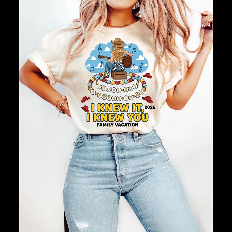 May include: A cream-colored short-sleeve t-shirt featuring a graphic of a cowgirl playing guitar on a crate. Below, a beaded necklace design displays the text YOU'VE GOT A FREND IN ME. Additional text reads I KNEW IT, I KNEW YOU 2026 FAMILY VACATION.