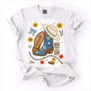 May include: A white t-shirt featuring a graphic print of a blue cowboy boot with floral patterns, a beige cowboy hat, sunflowers, a blue gingham bow, a stack of blocks with the text T 13 S, and a beaded string with the text I KNEW IT Y I KNEW N Y OU.
