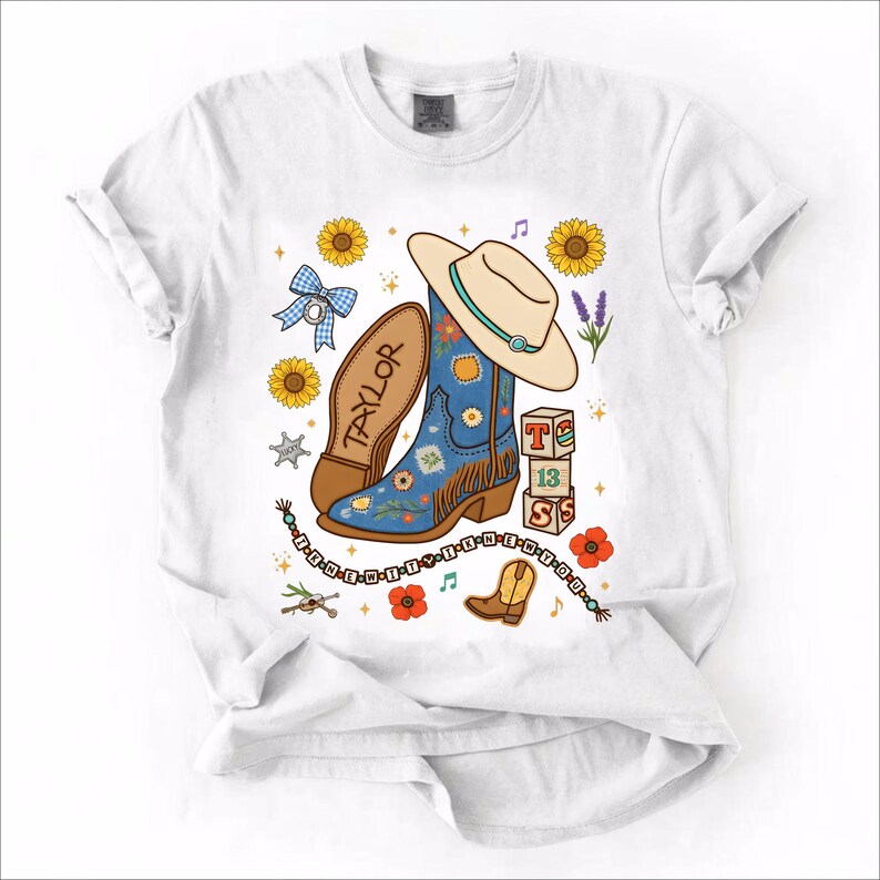 May include: A white t-shirt featuring a graphic print of a blue cowboy boot with floral patterns, a beige cowboy hat, sunflowers, a blue gingham bow, a stack of blocks with the text T 13 S, and a beaded string with the text I KNEW IT Y I KNEW N Y OU.