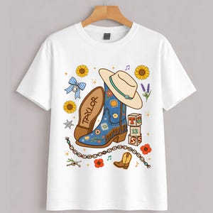 May include: A white short-sleeve t-shirt featuring a graphic print of a blue cowboy boot, a beige cowboy hat, sunflowers, a blue gingham bow, alphabet blocks spelling T-13-S, and a beaded string with the text I-R-N-E-W-I-T-T-Y-I-K-N-E-W-Y-O-U.