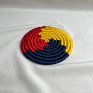 May include: A circular coaster featuring a coiled design with alternating segments of vibrant red, bright yellow, and deep navy blue thread. The tightly wound construction creates a smooth, flat surface with a distinct spiral pattern radiating from the centre.