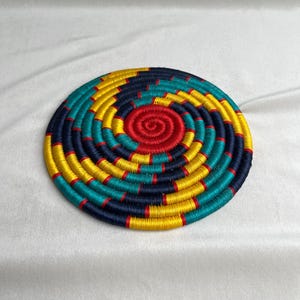 May include: A circular coaster featuring a vibrant spiral pattern created with tightly wound thread. The design consists of alternating segments of bright yellow, deep navy blue, teal, and red, radiating outwards from a solid red centre point.