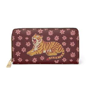 Puede incluir: Cartera burdeos con cremallera con una ilustración de tigre de dibujos animados en naranja y crema. La cartera está decorada con motivos florales rosas y tiene una cremallera y un tirador dorados.
