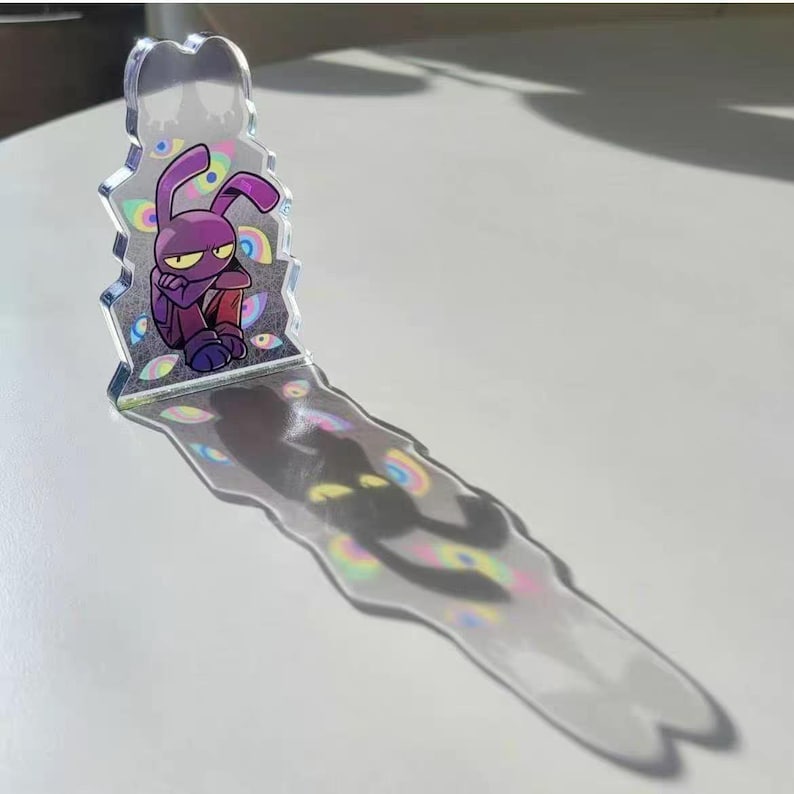 Op de afbeelding: Een helder acryl figuur met een paars cartoon konijn met een verdrietige uitdrukking. Het konijn zit met gekruiste armen. Het figuur werpt een lange, kleurrijke schaduw met oogvormige ontwerpen.
