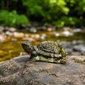 May include: A small, translucent green glass turtle figurine with a detailed, textured shell pattern and a smooth, rounded head. The sculpture features subtle dark green and brown mottled markings throughout its body, capturing a realistic turtle form.