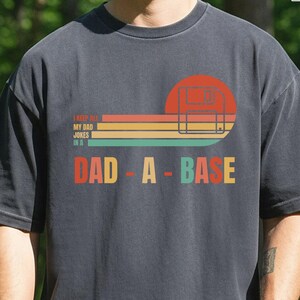 May include: Dark grey t-shirt with a retro design. The graphic features a red circle, horizontal stripes in orange, yellow, and green, and the words "DAD-A-BASE" in colourful letters. Above the text is the phrase "I KEEP ALL MY DAD JOKES IN A".