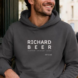 Peut inclure: Un sweat-shirt à capuche gris anthracite avec le texte blanc RICHARD BEER, le sous-titre UN SOURIRE - UNE MOUSSE - UN DATE et le texte plus petit REF CLUB imprimés sur la poitrine. Le vêtement possède une poche kangourou et des manches longues.