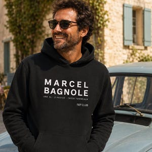 Peut inclure: Un sweat-shirt à capuche noir avec un texte blanc indiquant MARCEL BAGNOLE, UNE 4L, 2 PASTIS, TROIS TONNEAUX et REF CLUB. Ce vêtement possède des manches longues, une poche kangourou sur le devant et une capuche à cordon de serrage.