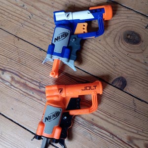 May include: Two toy NERF guns. One is blue, white, and orange, the other is orange and black. The blue gun has the word "NERF" printed on it. The orange gun has the word "JOLT" printed on it.