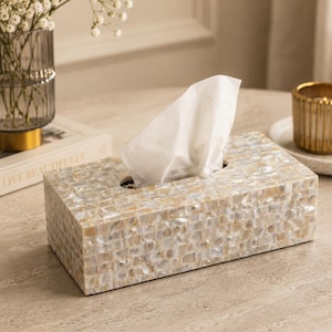 Puede incluir: Una caja para pañuelos rectangular que presenta un mosaico de azulejos de nácar iridiscente de color crema. La caja tiene una abertura ovalada central con un pañuelo blanco que sobresale por la parte superior, mostrando una superficie brillante.