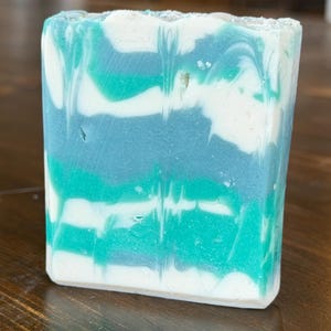 May include: A rectangular bar of handmade soap with a layered design. The soap features swirls of white, teal, and shades of blue. The soap is sitting on a wooden surface.