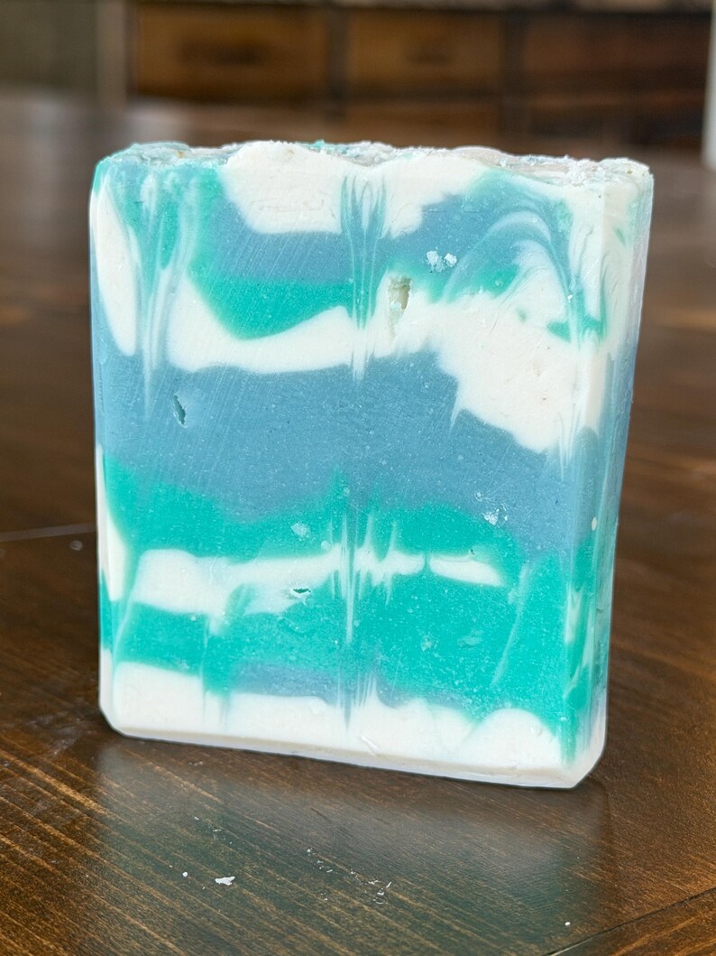 May include: A rectangular bar of handmade soap with a layered design. The soap features swirls of white, teal, and shades of blue. The soap is sitting on a wooden surface.