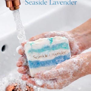 May include: A bar of soap with blue and white stripes is held in soapy hands under running water. The soap is labeled "Seaside Lavender". The faucet is copper-colored.