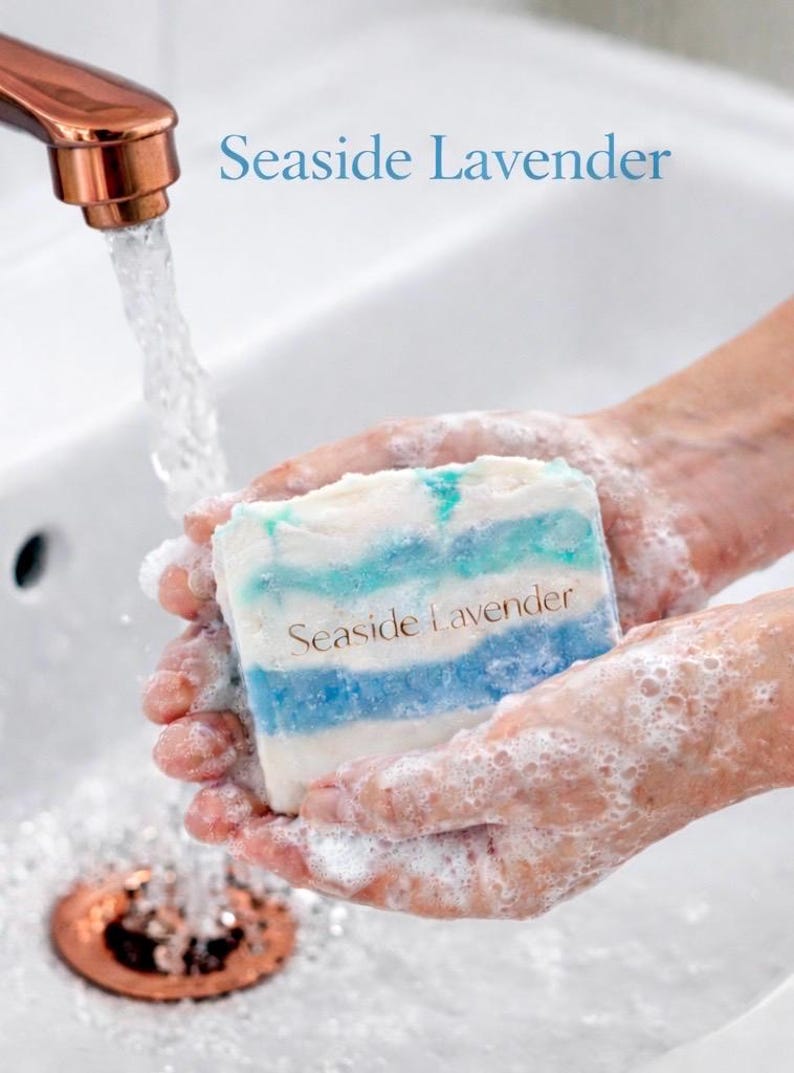 May include: A bar of soap with blue and white stripes is held in soapy hands under running water. The soap is labeled "Seaside Lavender". The faucet is copper-colored.