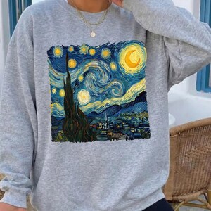 Puede incluir: Una sudadera gris con un estampado de la pintura "La noche estrellada" de Van Gogh. La obra de arte representa un cielo nocturno arremolinado con una luna y estrellas sobre un pueblo. La sudadera es de manga larga y tiene cuello redondo.