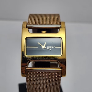 Op de afbeelding: Goudkleurig DKNY-horloge met een rechthoekige zwarte wijzerplaat en gouden wijzers. Het horloge heeft een mesh band en een goudkleurige kast. Het horloge is een stijlvol accessoire.