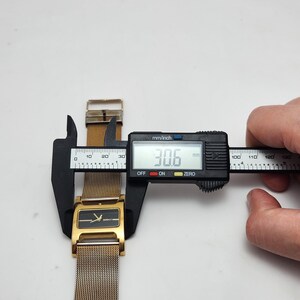 May include: A gold-toned watch with a mesh band is measured with a digital caliper. The watch face is rectangular with a black background and gold accents. The caliper displays a measurement of 30.6 mm.
