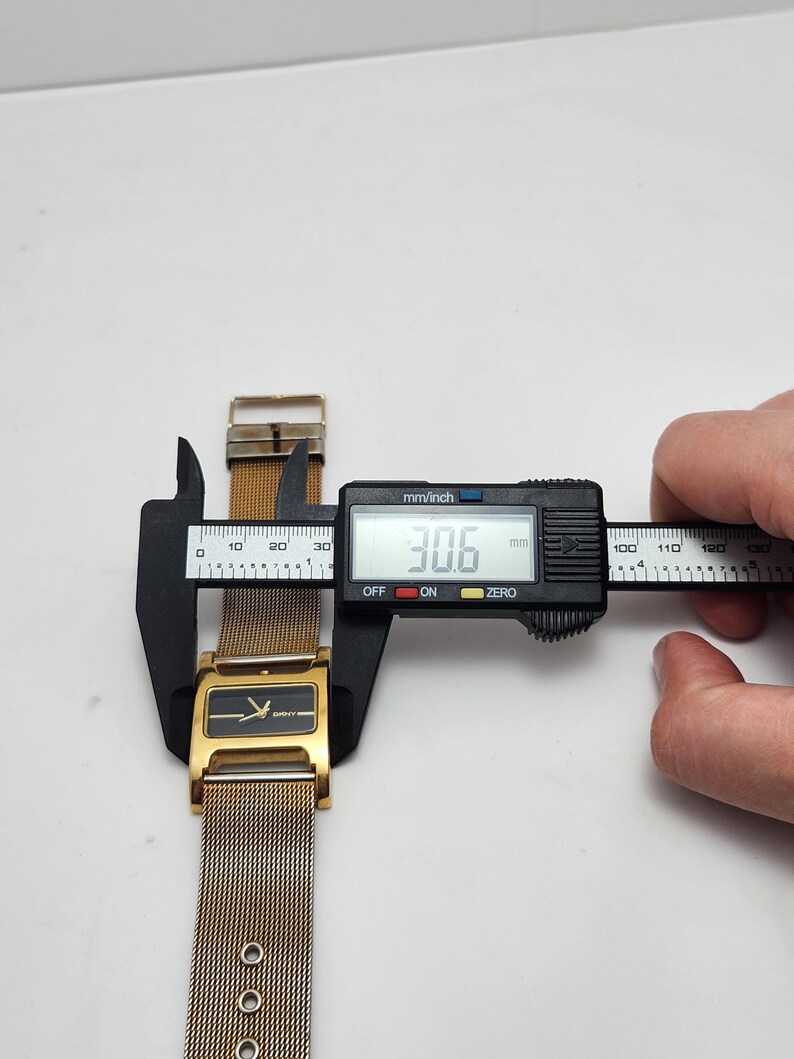 May include: A gold-toned watch with a mesh band is measured with a digital caliper. The watch face is rectangular with a black background and gold accents. The caliper displays a measurement of 30.6 mm.