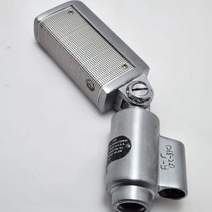 Puede incluir: Un micr&oacute;fono plateado vintage con una pantalla perforada y un cuerpo cil&iacute;ndrico. El micr&oacute;fono tiene un dise&ntilde;o direccional e incluye una etiqueta con texto. Es un equipo de audio cl&aacute;sico.