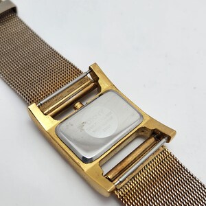 May include: A gold-toned DKNY watch with a mesh band. The watch face is rectangular with a silver backing. The band is a fine mesh of gold-colored metal. The watch is water resistant.